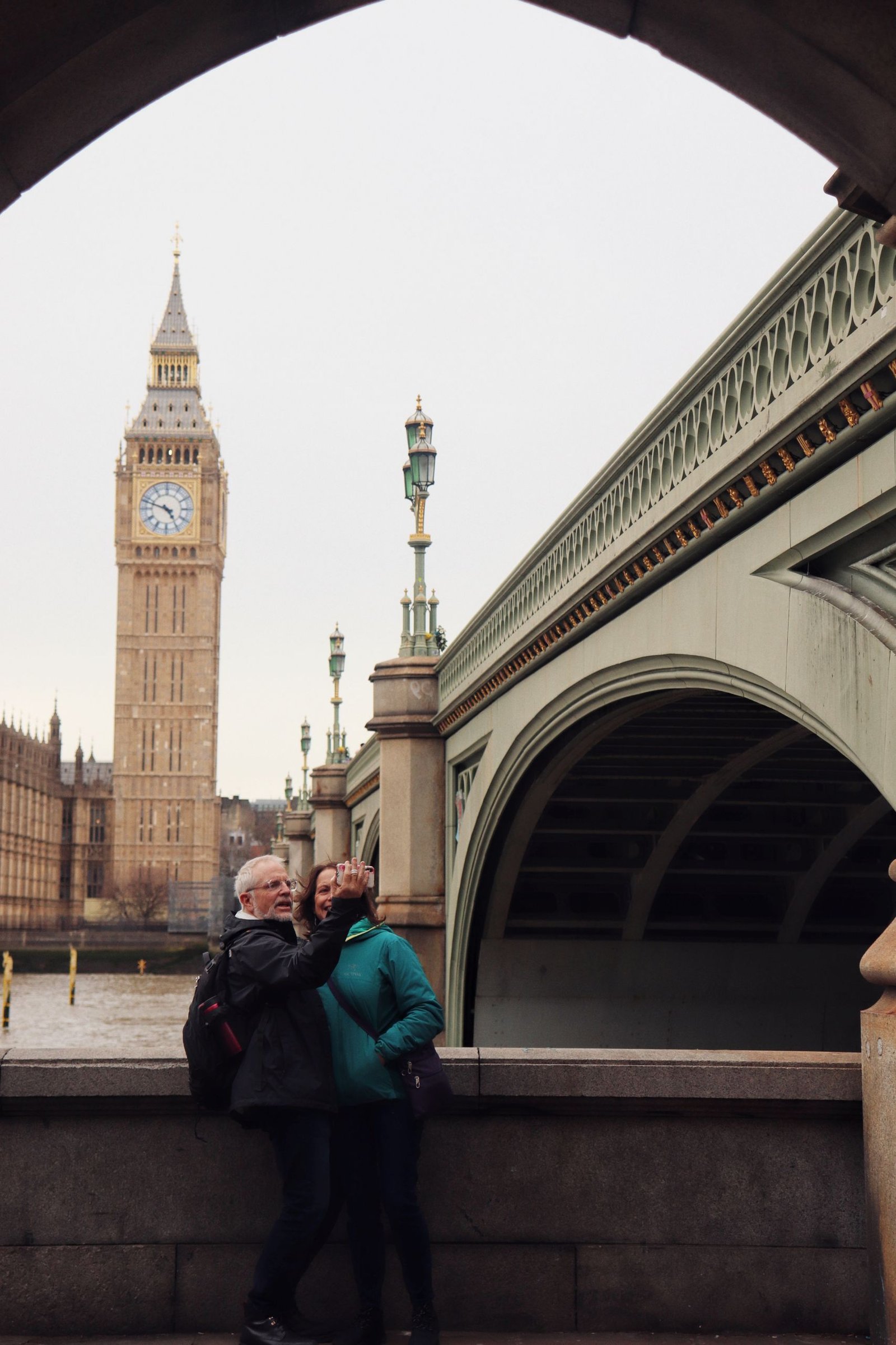A couple poses for a selfie near Big Ben and a bridge in London.